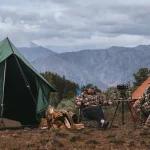 Hunters relaxing at a well-organized backcountry hunting camp with canvas tents, secured gear, and a mountain landscape in bear country.