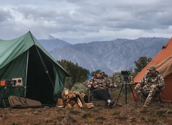 Hunters relaxing at a well-organized backcountry hunting camp with canvas tents, secured gear, and a mountain landscape in bear country.