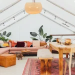 Spacious luxury glamping tent interior with cozy seating, wooden dining furniture, rugs, plants, and warm ambient lighting.