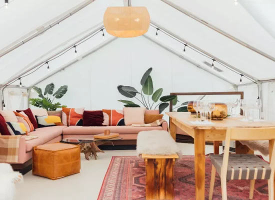 Spacious luxury glamping tent interior with cozy seating, wooden dining furniture, rugs, plants, and warm ambient lighting.