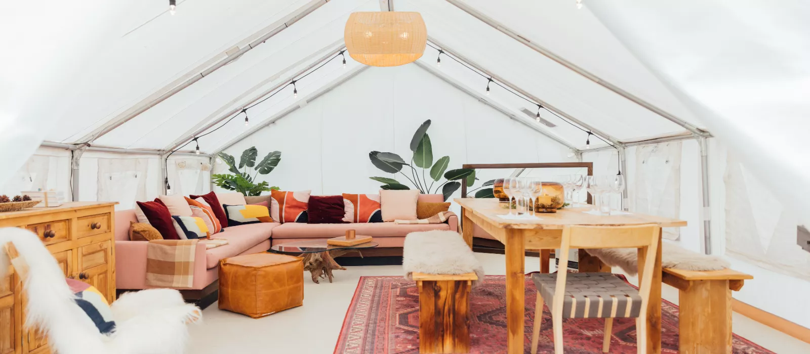 Spacious luxury glamping tent interior with cozy seating, wooden dining furniture, rugs, plants, and warm ambient lighting.