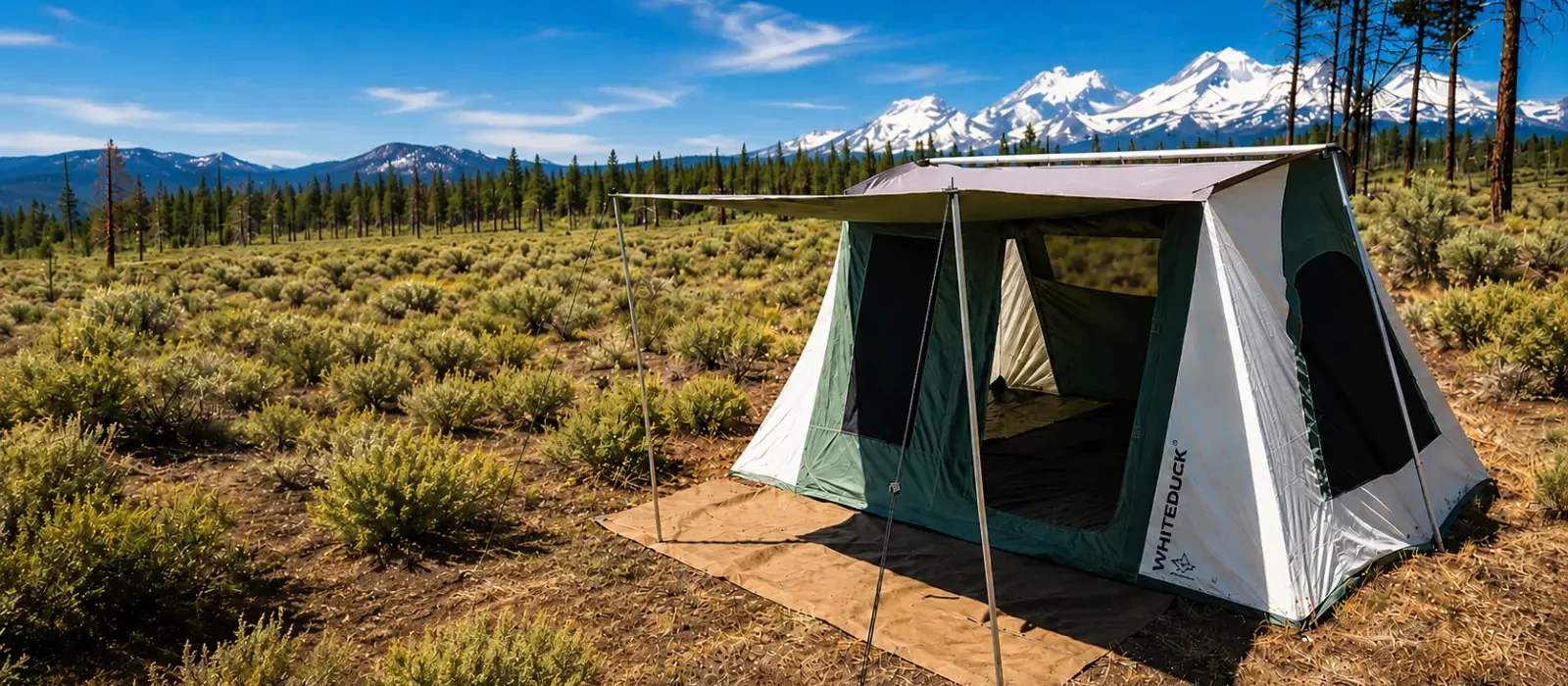 White Duck Prota Cabin Tent set up at a mountain campsite surrounded by pine trees and snow-capped mountains