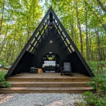Black A-frame cabin with a wooden deck nestled among green trees in a forest glamping setting.