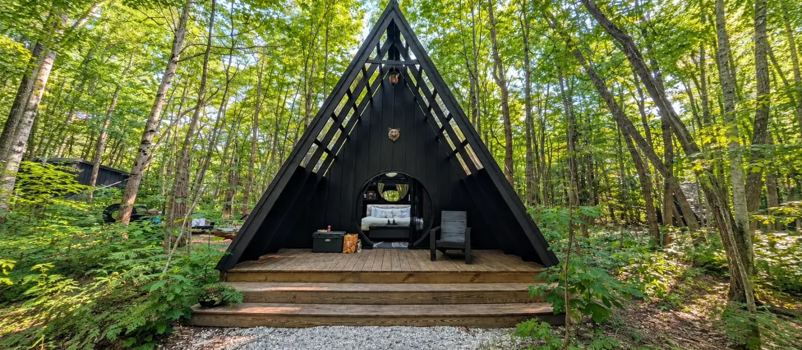 Black A-frame cabin with a wooden deck nestled among green trees in a forest glamping setting.