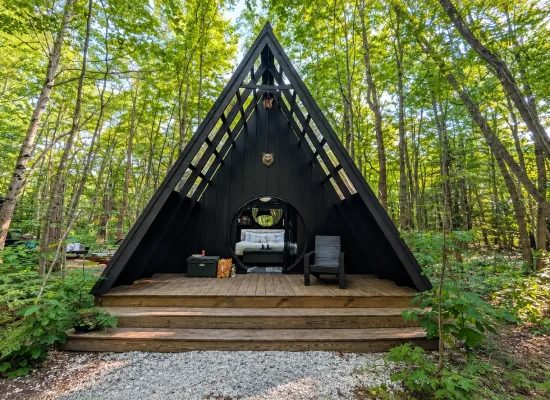 Black A-frame cabin with a wooden deck nestled among green trees in a forest glamping setting.