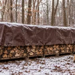 Firewood stack covered with a brown poly tarp to protect logs from snow and winter weather, with a canvas log carrier beside it.