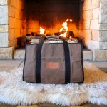 White Duck canvas log carrier in brown with black handles, filled with birch logs and set on a sheepskin rug in front of a lit stone fireplace