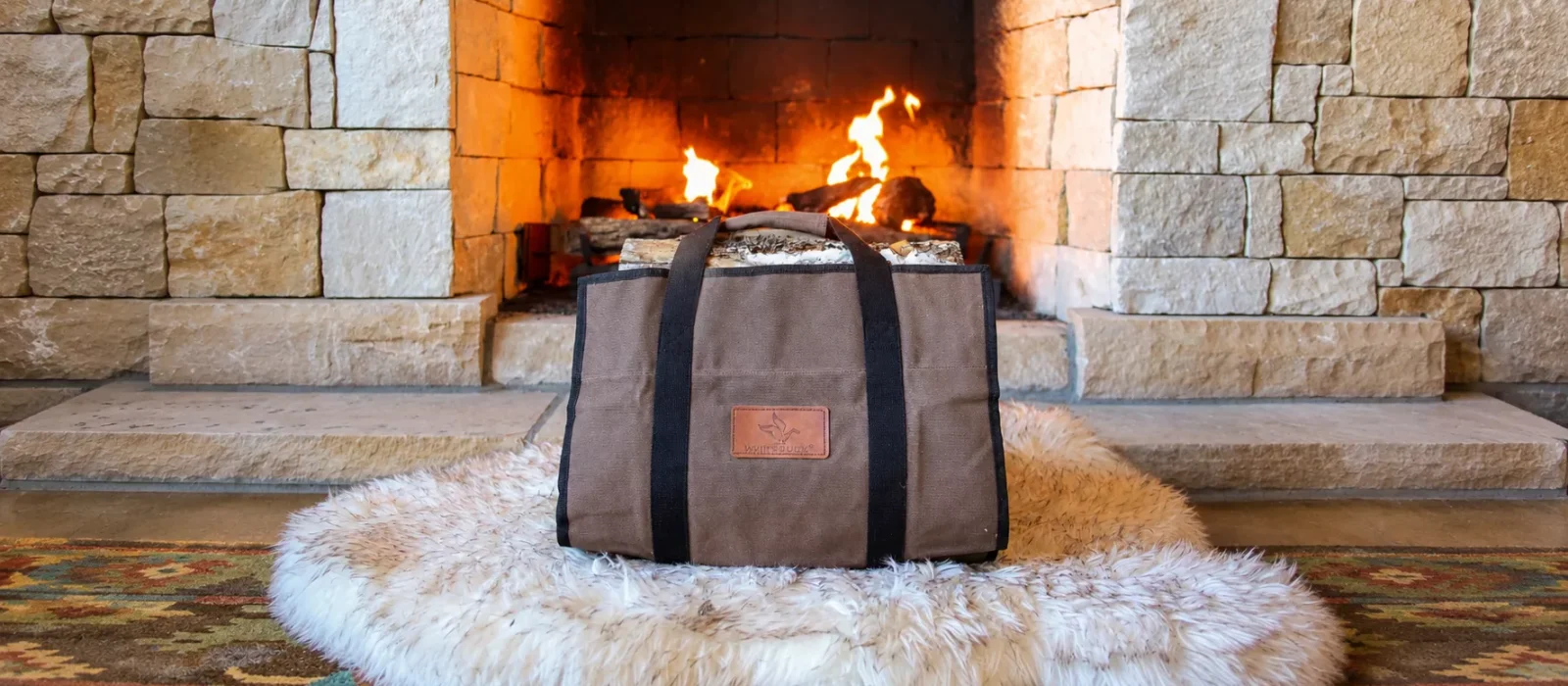 White Duck canvas log carrier in brown with black handles, filled with birch logs and set on a sheepskin rug in front of a lit stone fireplace