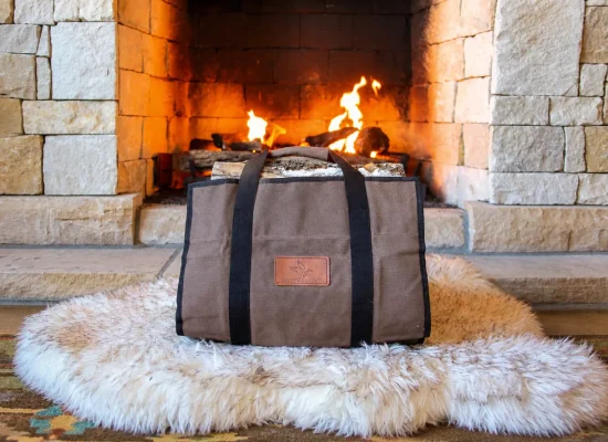 White Duck canvas log carrier in brown with black handles, filled with birch logs and set on a sheepskin rug in front of a lit stone fireplace
